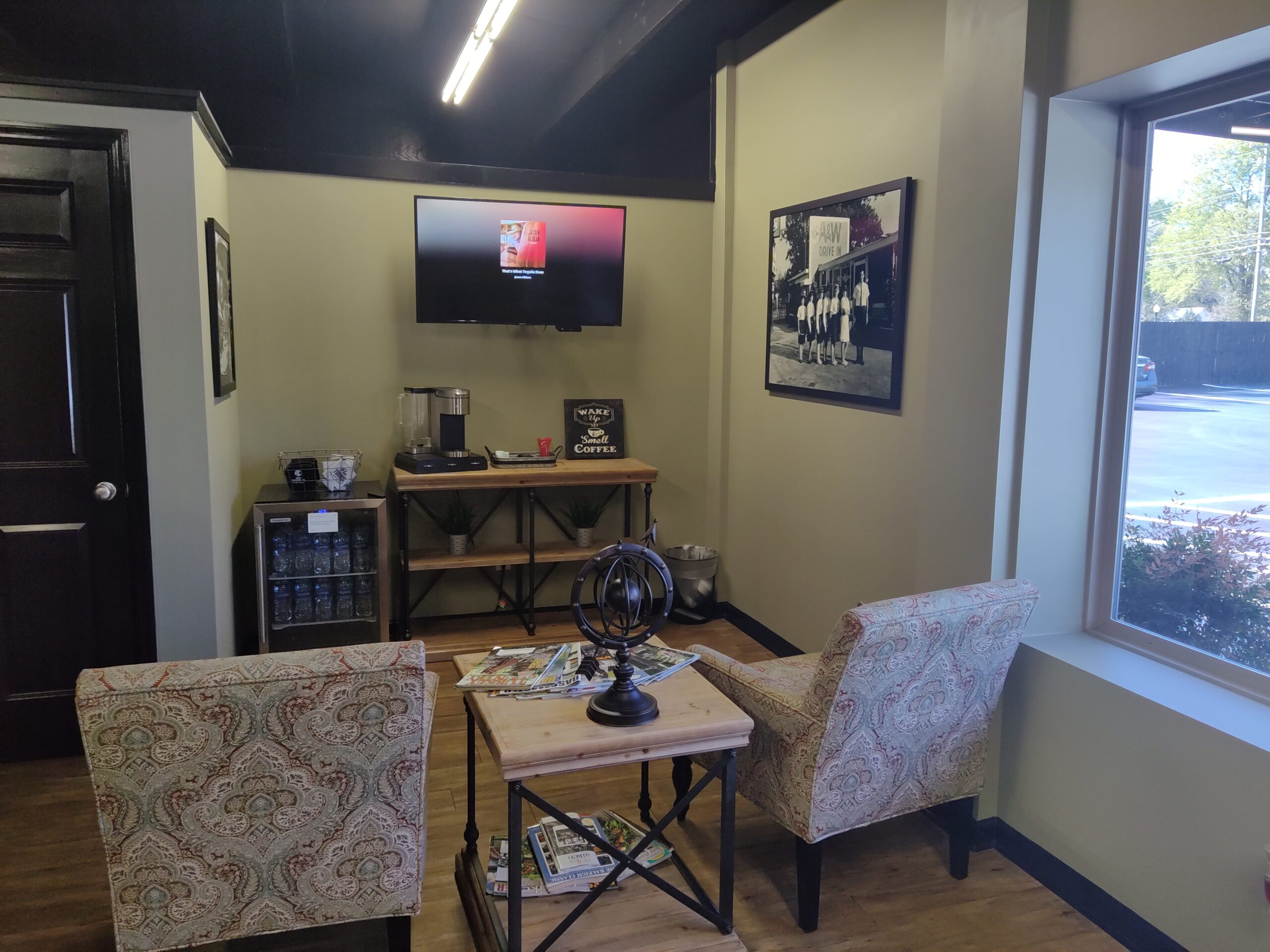 Photo features sitting area at Aiken Collision. Coffee bar, TV, 2 chairs, table and a refrigerator of refreshments with logo drink covers.